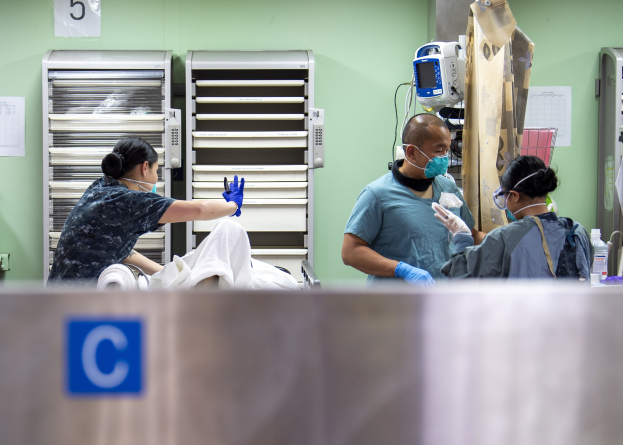 Drei medizinische Mitarbeiter in Kitteln und Masken versorgen einen Patienten auf einem mit einem weißen Tuch bedeckten Krankenhausbett, mit medizinischen Versorgungsmitteln auf einem nahen Tisch und Geräten im Hintergrund.