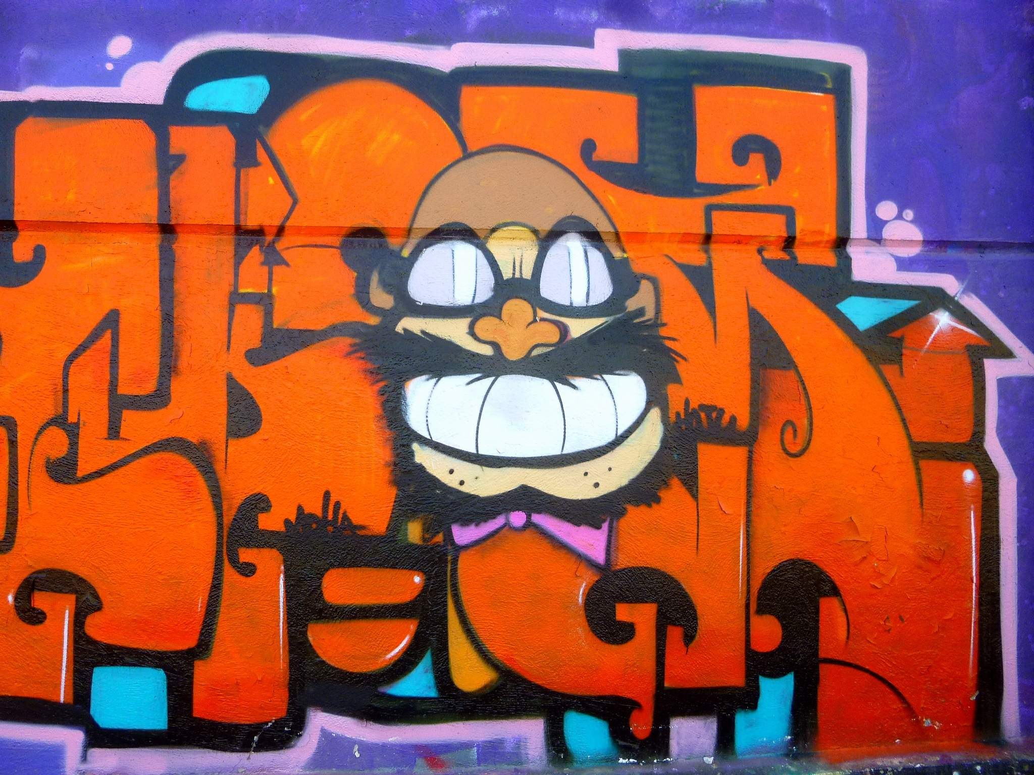 Graffiti-Gemälde eines Cartoon-Charakters mit großen Augen, einem breiten Lächeln und einem verschmitzten Gesichtsausdruck an der Seite eines Gebäudes, mit leuchtenden Farben und auffälliger Begleittext.