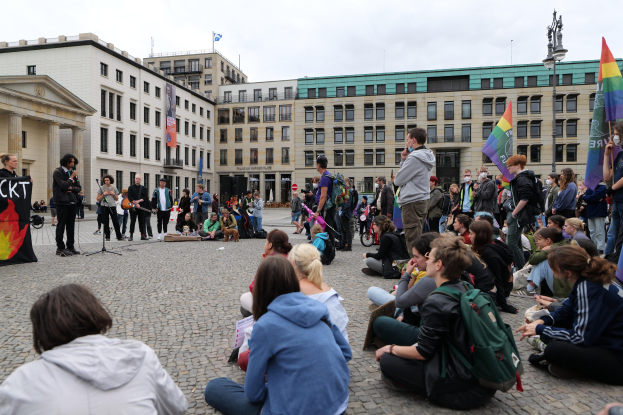 Eine Gruppe von Menschen, die auf dem Boden sitzen und vor einer Menge mit Fahnen und Plakaten sprechen, mit einer Statue und Gebäuden im Hintergrund während einer Homo-Protest in Berlin, Deutschland.