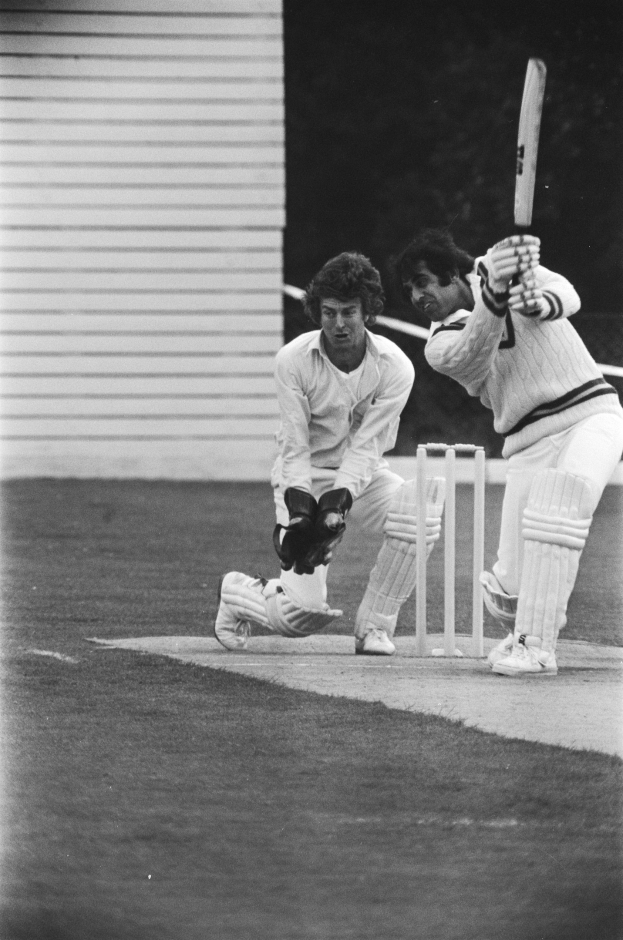 Zwei Männer spielen Cricket auf einem Feld, einer hält einen Schläger vor einem Wicket in Schwarz-Weiß.