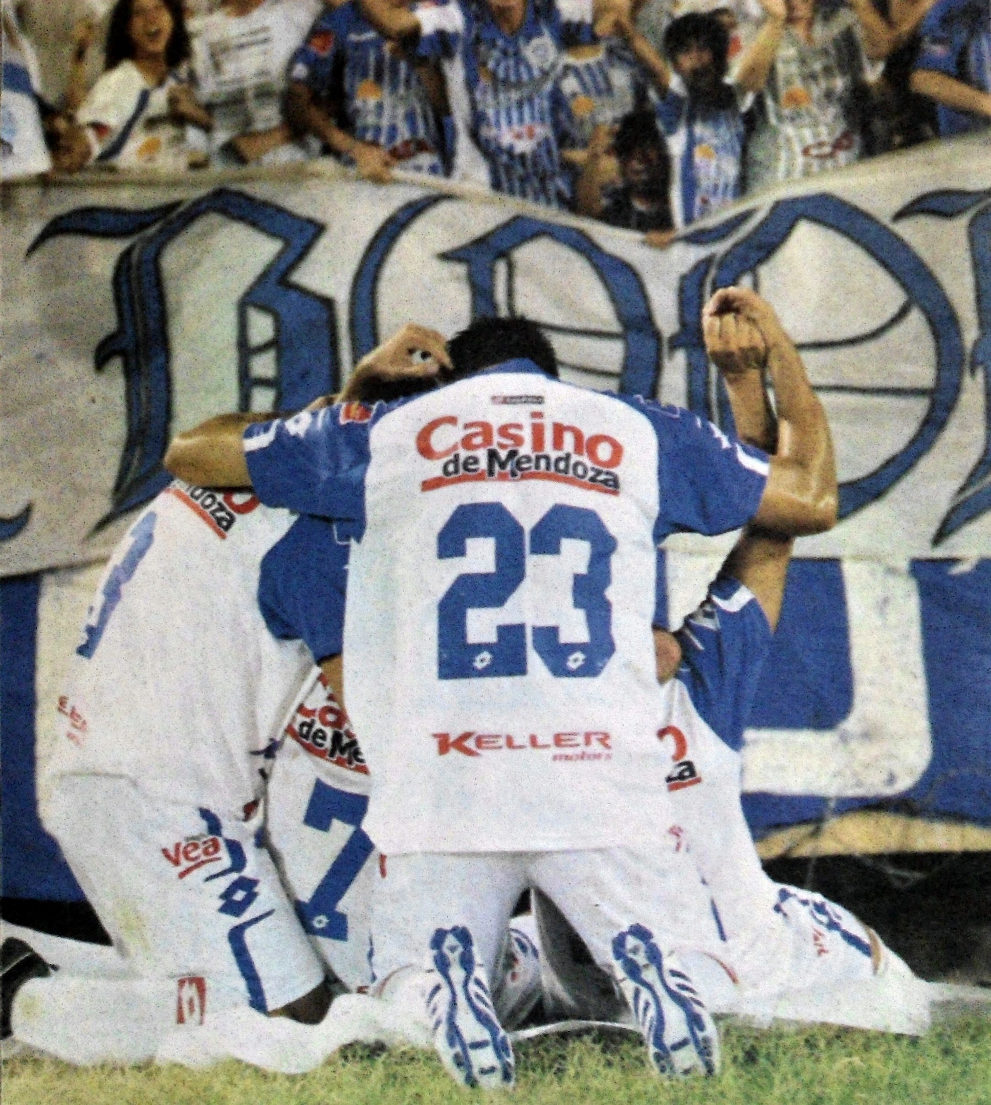 Gruppe von Fußballspielern, die vor jubelnden Fans auf dem Boden feiern, mit einem Banner im Hintergrund.