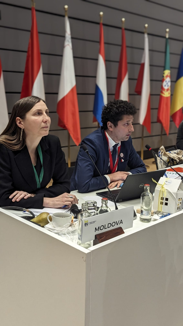 Gruppe von Menschen an einem Tisch mit Laptops, Flaschen, Tassen, Namensschildern und Mikrofonen während einer Europäischer Ratssitzung über Moldau mit sichtbaren Flaggen im Hintergrund.