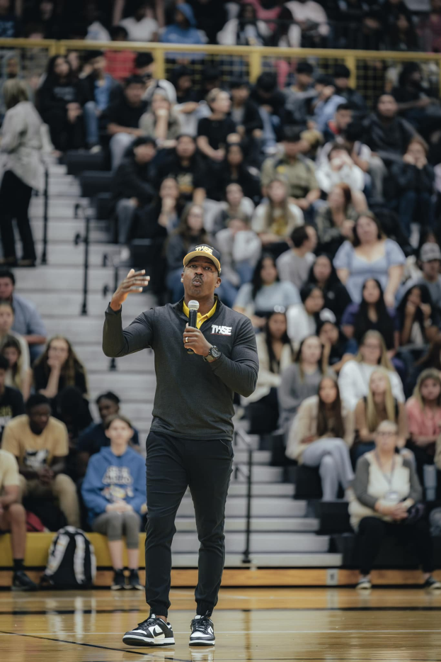Ein Mann in einer Mütze und mit einem Mikrofon in der Hand steht auf einem Basketballfeld, mit einer sitzenden Menge und Taschen dahinter, was auf ein Basketballspiel hinweist, bei dem er wahrscheinlich der Cheftrainer ist.