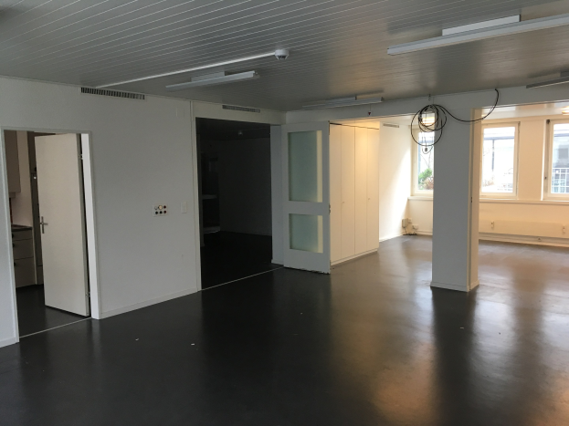 Leeres Zimmer mit schwarzen Böden, weißen Wänden, elektrischen Lampen an der Decke, sichtbaren Kabeln, Türen, Schränken, Fenstern und einer Außenansicht von Gebäuden und Bäumen, gelegen in einem Büroraum zur Miete in Amsterdam.