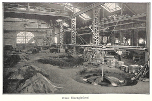 Black and white photo of a factory with workers operating machinery, surrounded by metal rods and poles, with text at the bottom reading "New Eisenglebeerei, the first factory in Germany".