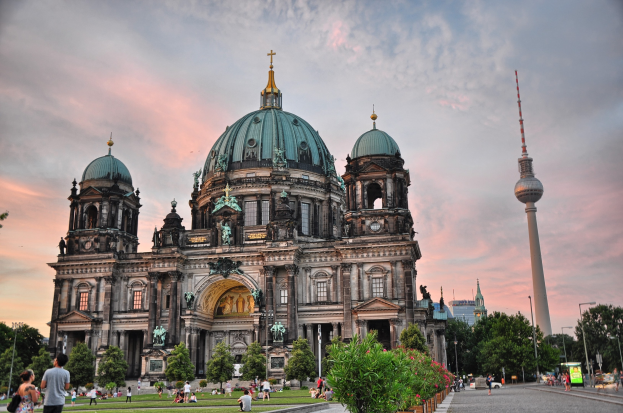 Große Kathedrale mit Kuppel, Säulen, Bögen und Statuen, umgeben von Grün, Menschen, Fahrzeugen und Straßenelementen unter einem bewölkten Himmel.