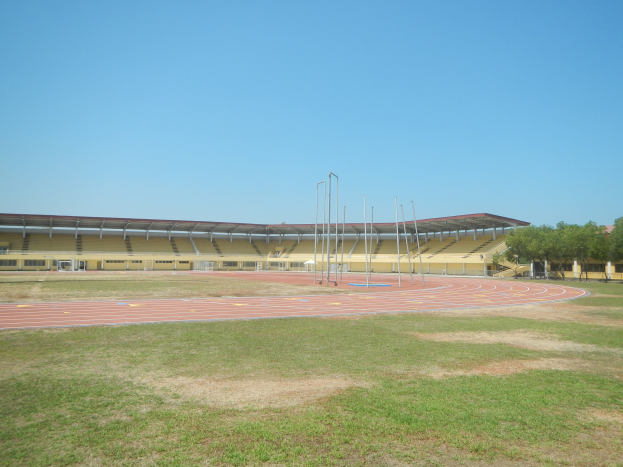 Großes Stadion mit einer zentralen Laufbahn, umgeben von grünem Gras und Bäumen auf der rechten Seite, unter einem klaren blauen Himmel mit Pfosten im Hintergrund.