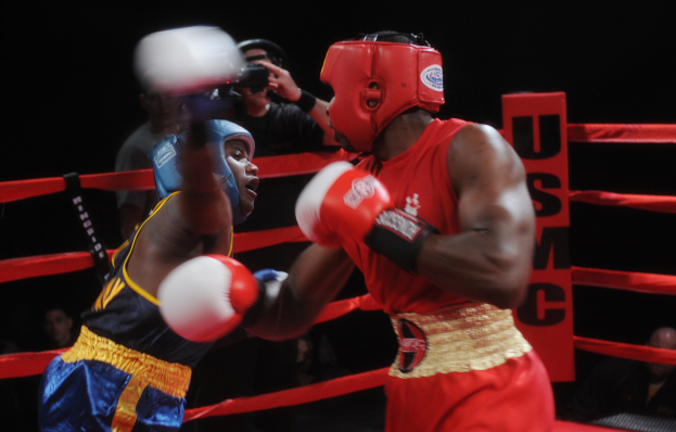 Zwei Boxer im Ring während eines Kampfes, mit einem Fotografen und Zuschauern im Hintergrund bei dramatischer Beleuchtung.