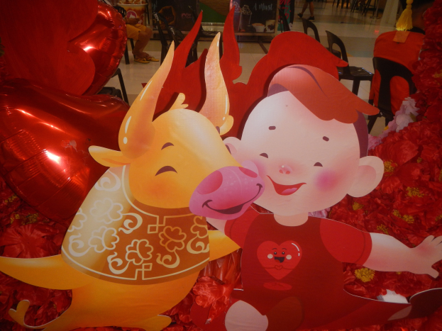 Eine festliche Szene des chinesischen Neujahrsfestes mit einem Cartoon-Jungen und einem Schwein, umgeben von roten Blumen und Ballons, mit Menschen und dekorierten Brettern im Hintergrund.