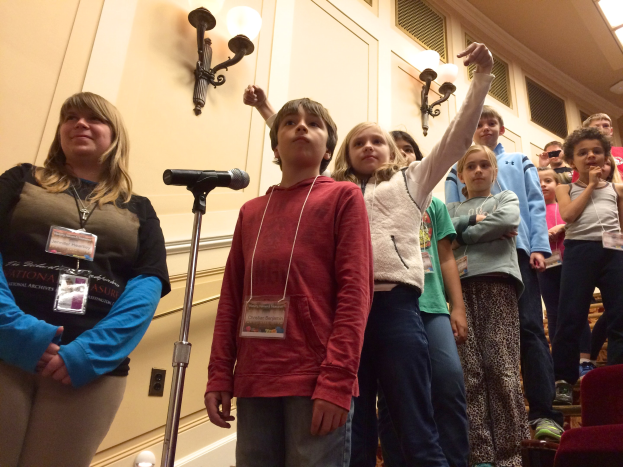 Eine Gruppe von Kindern singt an einem Mikrofon mit einer Frau daneben, Stühle auf der rechten Seite und Wandlichter im Hintergrund.