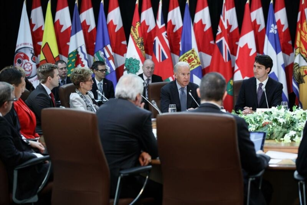 Gruppe von Menschen an einem Tisch mit Flaggen im Hintergrund, die in eine Diskussion mit Mikrofonen, Gläsern, Blumen, Papieren und anderen Gegenständen auf dem Tisch vertieft sind, wahrscheinlich im Zusammenhang mit einem Kanada-Kanada-Handelsabkommen.