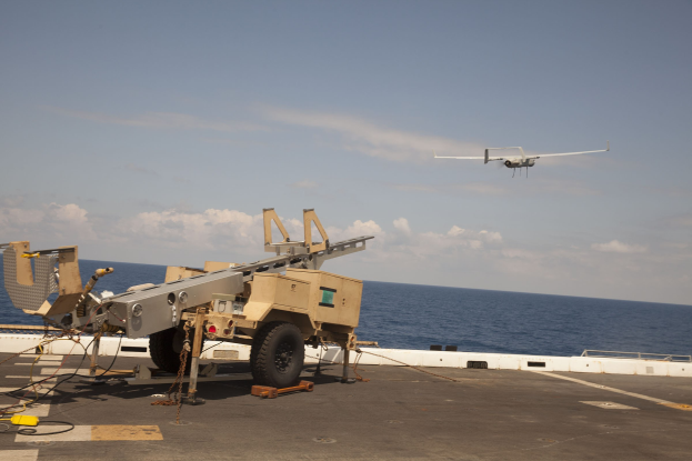Militärfahrzeug auf dem Deck eines Flugzeugträgers mit einer Drohne darüber, Wolken am Himmel und Wasser im Hintergrund.
