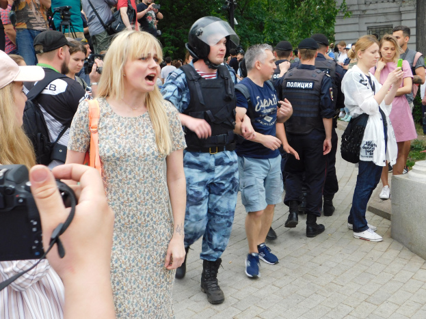 A group of people stands in front of a protesting crowd, some wearing caps and carrying bags or cameras, with trees and a building in the background.