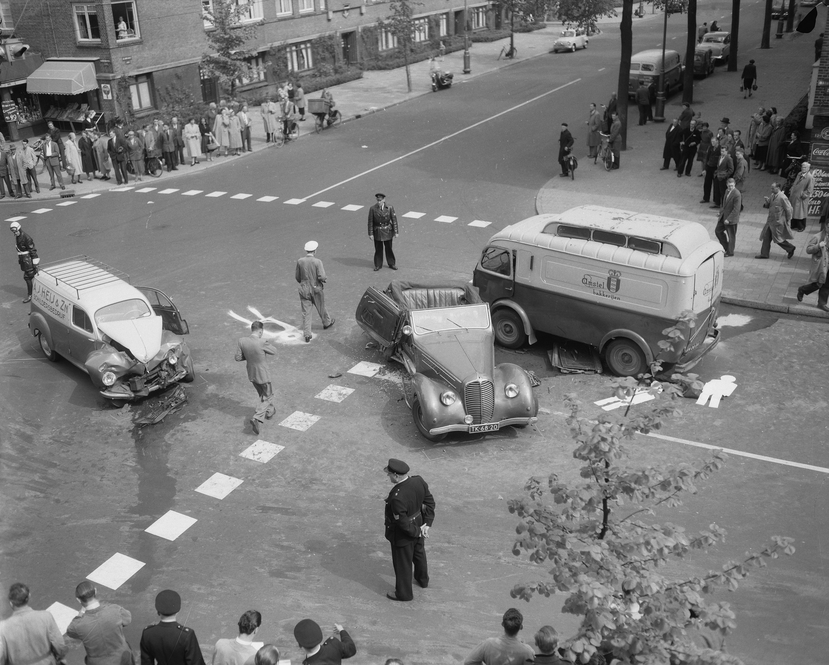 Schwarze und weiße Szenerie eines Autounfalls an einer Straßenecke mit mehreren Fahrzeugen, darunter ein Lieferwagen, und einer Gruppe von Menschen in der Nähe, mit Gebäuden, Bäumen, Pfosten und Texttafeln im Hintergrund.