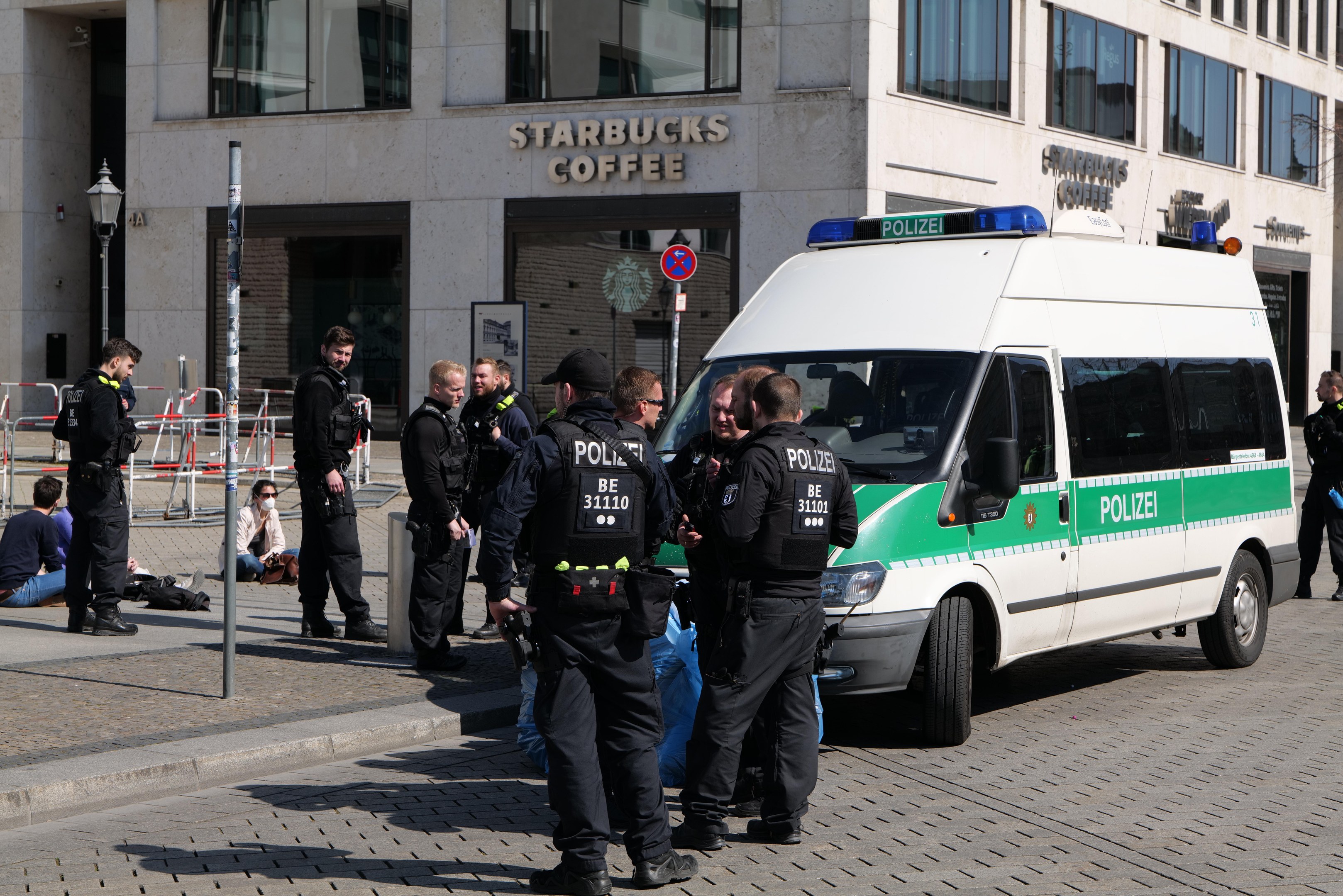 Polizisten vor einem Starbucks-Café mit einem Van auf der rechten Seite und Menschen auf der linken Seite, mit einem Gebäude, Schild, Laterne und Zaun im Hintergrund.