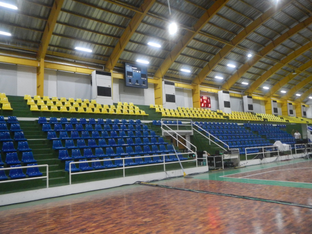 Großer Hallensportplatz mit Holzboden, blauen und gelben Sitzplätzen, Geländern, Treppen zum Sitzbereich, Wandbildschirmen, Deckenleuchten und einigen Menschen.