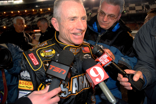 Ein Mann in einer schwarzen Jacke spricht mit Reportern vor einem hell erleuchteten Stadion voller Menschen, einige halten Mikrofone und einer eine Kamera.