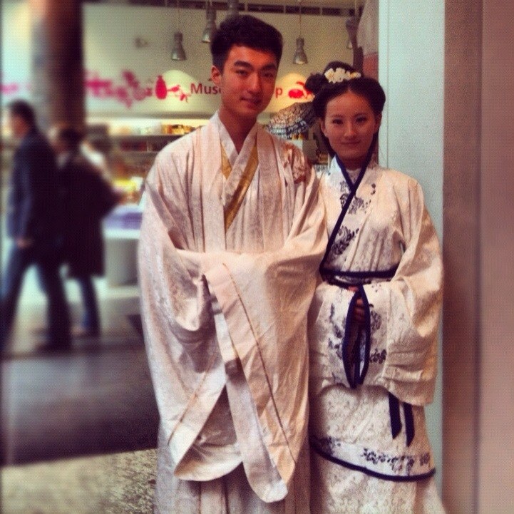 Zwei Menschen in traditioneller koreanischer Kleidung stehen lachend nebeneinander, mit unscharfen Figuren und Lichtern im Hintergrund.