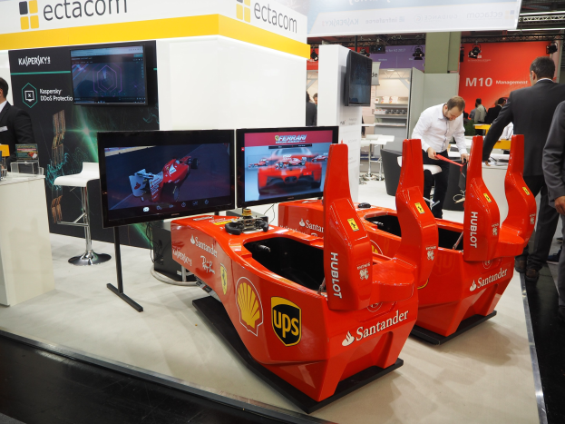 Eine Gruppe von Menschen um einen roten Ferrari F1 auf einem Messestand herumstehend, umgeben von Fernsehern, Möbeln und Texttafeln in einem hell erleuchteten Veranstaltungsort.