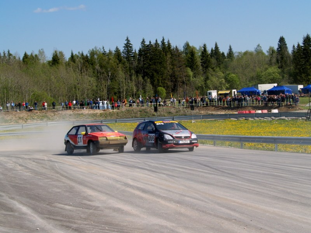 Zwei Autos fahren auf einer Schotterstraße an einer Menschenmenge vorbei, getrennt durch ein Geländer, mit gelben Blumen auf der rechten Seite und Bäumen im Hintergrund unter einem klaren blauen Himmel.