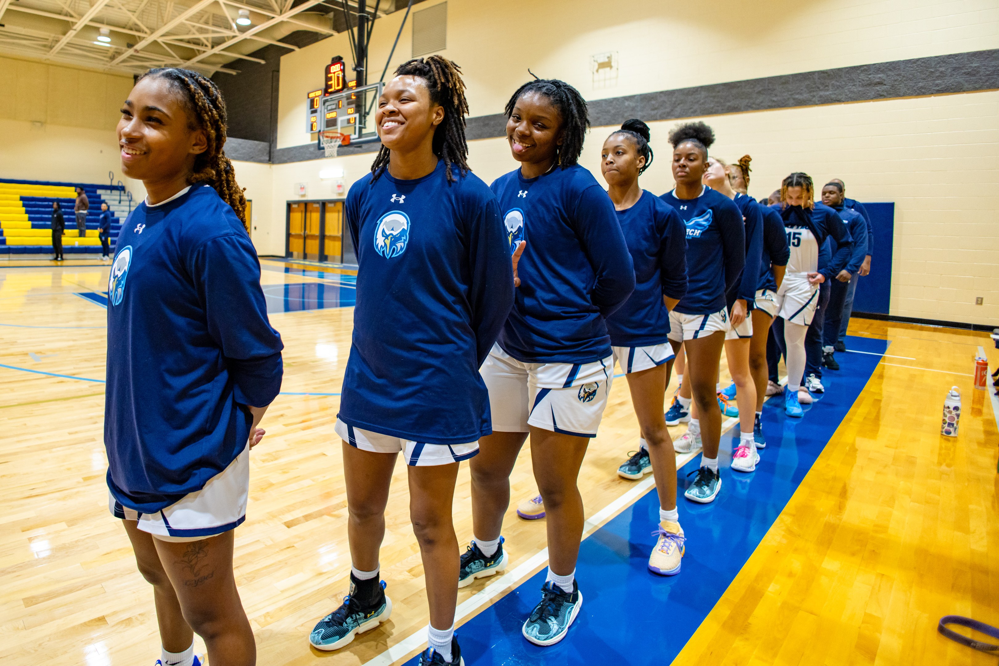 Eine Gruppe junger Frauen in blauen T-Shirts, die auf einem Basketballfeld lächeln, mit Flaschen und Gegenständen auf dem Boden, Menschen, die auf Treppen im Hintergrund sitzen, und einem Basketballkorb unter Deckenlampen.