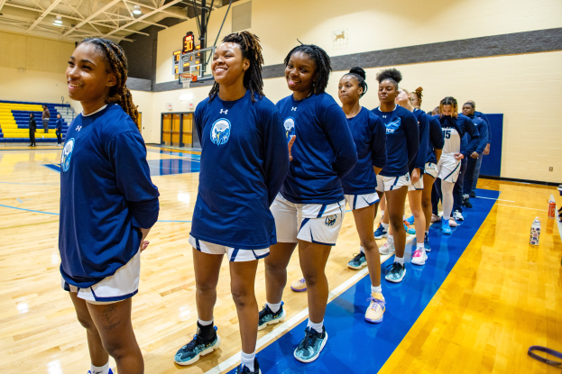 Eine Gruppe junger Frauen in blauen T-Shirts, die auf einem Basketballfeld lächeln, mit Flaschen und Gegenständen auf dem Boden, Menschen, die auf Treppen im Hintergrund sitzen, und einem Basketballkorb unter Deckenlampen.