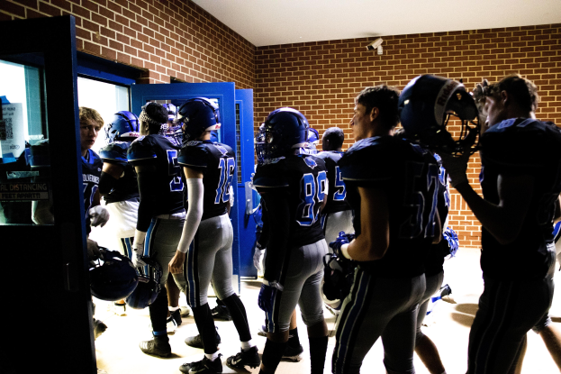 Gruppe von Fussballspielern in Uniformen und Helmen, die in einer Umkleidekabine mit sichtbaren Wänden und einer Tür auf der linken Seite stehen.