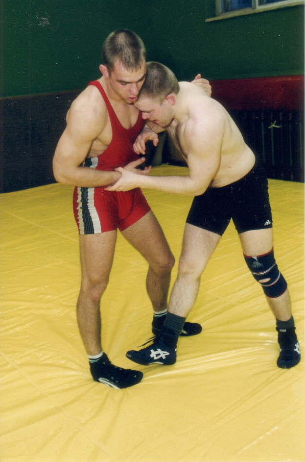 Zwei Männer ringen auf einer gelben Matte in einem Fitnessstudio, Arme fest ineinander verschränkt.