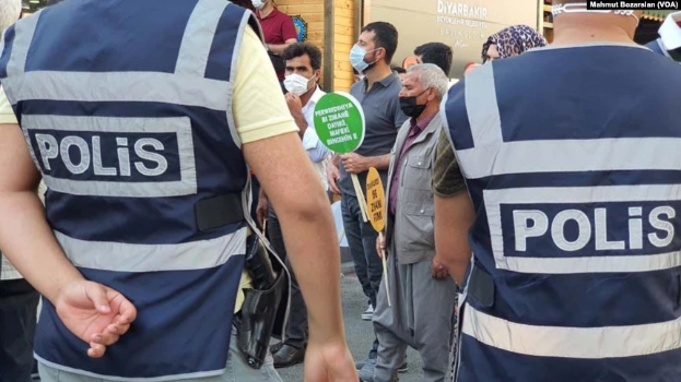 Eine Gruppe von Polizisten steht vor einer Menge mit Maskenschilden, mit einem verschlossenen Gebäude und Istanbul-Wasserzeichen im Hintergrund.