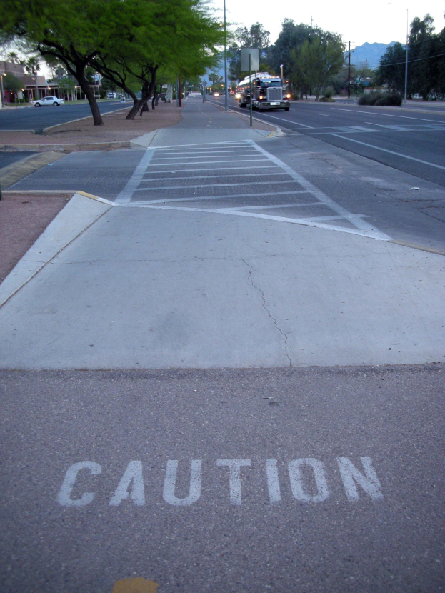 Eine Stadtstraße mit fahrenden Fahrzeugen, ein Gehweg mit einem Warnschild in der Nähe eines Baumes, Strommasten, Straßenlaternen, Bauplanken und Pflanzen, flankiert von Gebäuden und Bergen unter einem klaren blauen Himmel.