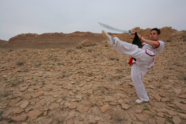 Ein Mann in einem weißen Karate-Anzug trainiert mit einem Schwert in einer Wüstenlandschaft mit steinigem Boden, Hügeln und einem klaren blauen Himmel.