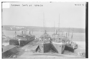 Ein Schwarz-Weiß-Foto mehrerer deutscher Schiffe, die an einem Dock mit Pfählen, Seilen und anderen Gegenständen vertäut sind, mit Bäumen und einem klaren Himmel im Hintergrund und Text oben auf dem Bild.