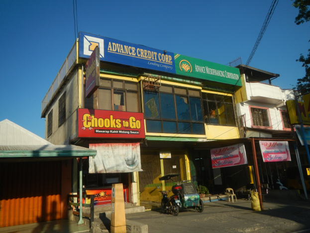 Ein Geschäftsgebäude mit einer Tafel, auf der "Advance Credit Corp" steht, neben einer vielbefahrenen Straße mit Fahrzeugen, Werbetafeln, Strommasten, Kabeln, Bäumen und einem klaren blauen Himmel.