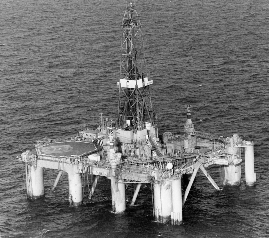 Schwarzes und weißes Foto einer Ölplattform in der Mitte des Ozeans, umgeben von Wasser, mit einer Brücke mit Säulen und einem Turm im Hintergrund.