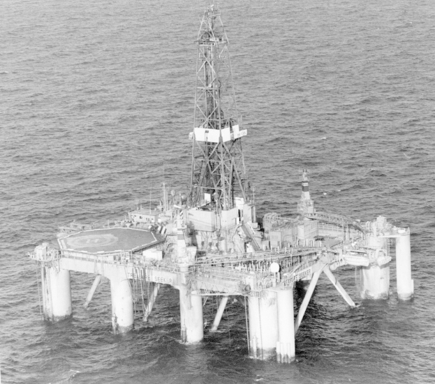Schwarzes und weißes Foto einer Ölplattform in der Mitte des Ozeans, umgeben von Wasser, mit einer Brücke mit Säulen und einem Turm im Hintergrund.