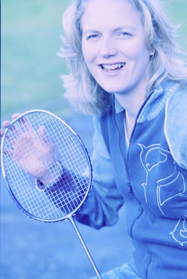 Eine Frau in einem weißen Hemd und blauen Shorts, die einen Badminton-Schläger hält und lächelt, mit einem Pferdeschwanz und konzentriertem Blick auf den Schläger.