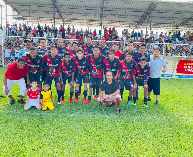 Gruppe von Männern, die auf einem Fußballfeld mit einem Siegesschild feiern, das den Text 'Fotos Ugarte U-17 vs U-18' trägt, unter einem bewölkten Himmel.