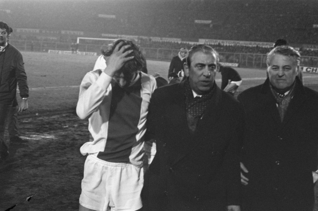 Schwarzes Bild einer Gruppe von Männern auf einem Fußballfeld mit einem Stadion im Hintergrund, die nach einem Sieg enttäuscht aussehen.