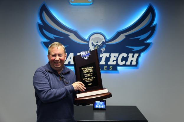 Ein Mann hält eine 'NCAA Fall National Championship'-Trophäe vor einer mit einem Logo verzierten Wand, wobei ein digitales Display auf einem Tisch dahinter sichtbar ist.
