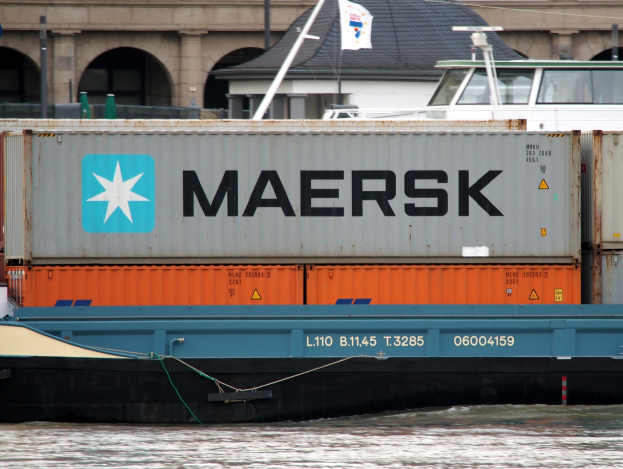 A barge carrying a large container with text travels down a river, with a building visible in the background.