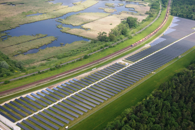 Aerial view of a solar farm with solar panels in a field, surrounded by trees, grass, plants, and water, with a train on a nearby railway track.