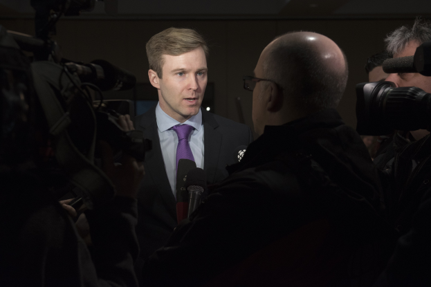 Ein Mann in Anzug und Krawatte mit Brille spricht zu Reportern mit zwei Personen mit Kameras auf jeder Seite und einem Mikrofon vor ihm.