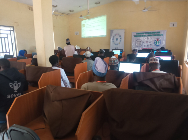 Eine Gruppe von Menschen, die an Computern in einem Seminarraum mit einem Bildschirm, einer Fahne, Deckenventilatoren und Fenstern sitzen.