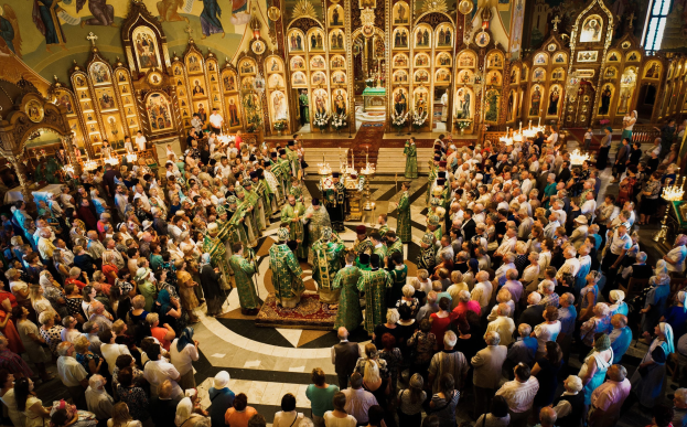 Eine große Gruppe von Menschen steht vor einer russisch-orthodoxen Kirche, einige halten Musikinstrumente, mit brennenden Kerzen auf Ständern, einem Teppichboden und gerahmten Fotos an den Wänden während einer religiösen Feier.