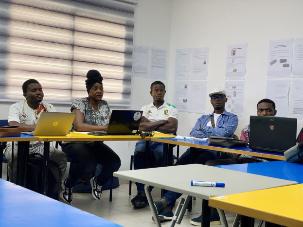 Gruppe von Menschen, die um einen Tisch sitzen und mit Laptops und anderen Gegenständen beschäftigt sind, in Diskussion mit Papieren an der Wand und Jalousien im Hintergrund.