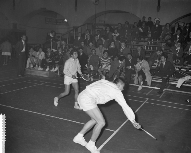 Zwei Männer spielen Badminton auf einem Platz mit Zuschauern im Hintergrund, beide halten Schläger.