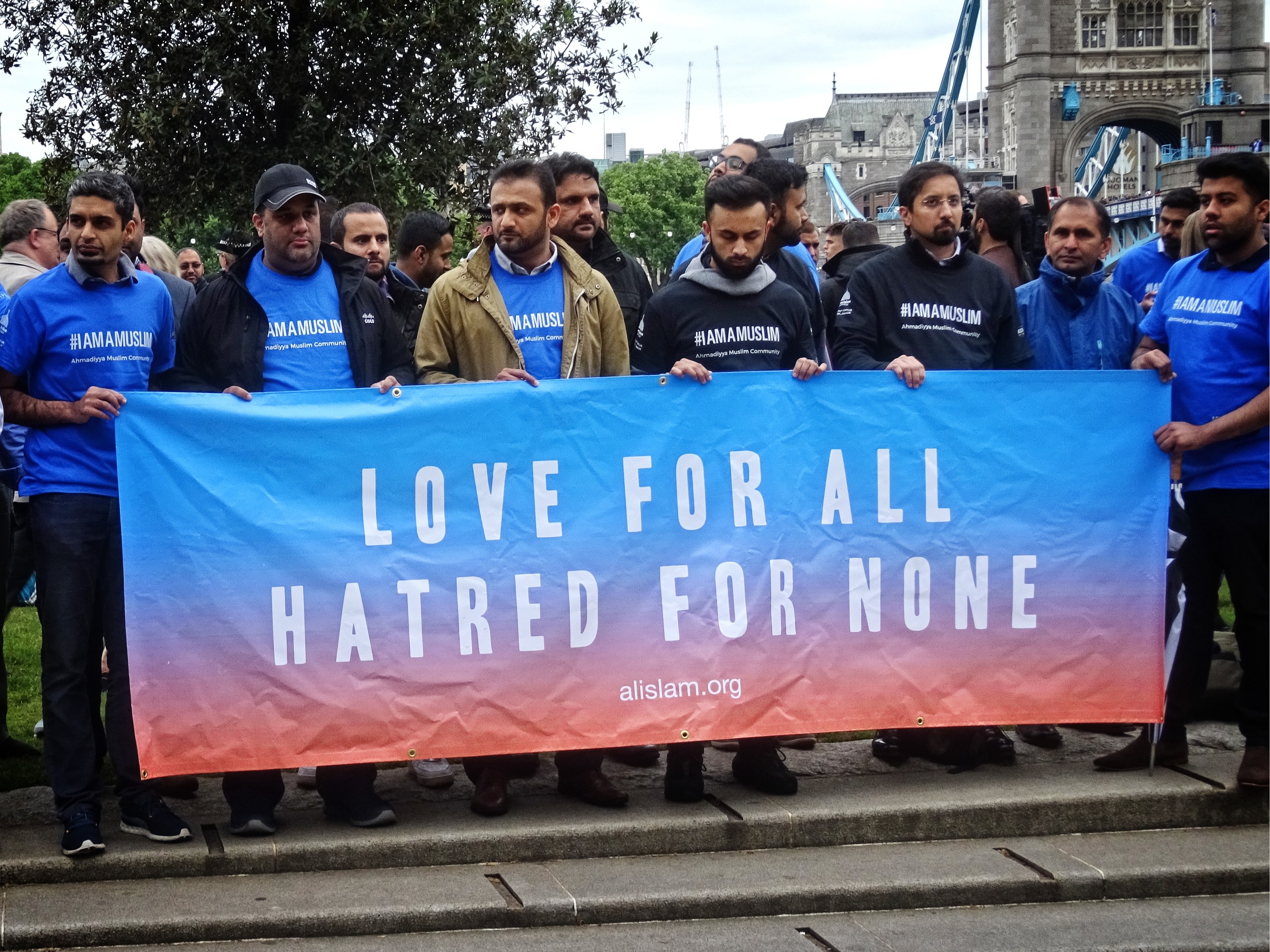 Eine Gruppe von Menschen hält ein Transparent mit der Aufschrift "Liebe für alle Hass für niemanden" vor einem Baum, Gebäuden und einem bewölkten Himmel.
