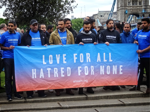 Eine Gruppe von Menschen hält ein Transparent mit der Aufschrift "Liebe für alle Hass für niemanden" vor einem Baum, Gebäuden und einem bewölkten Himmel.