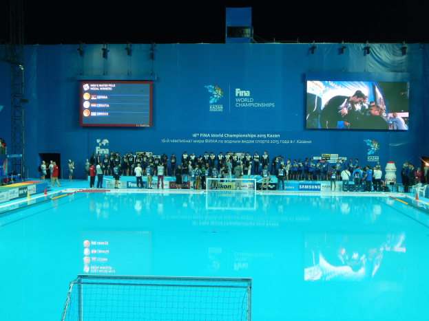Gruppe von Menschen, die um ein Hallenbad mit Netz am Boden stehen, mit einer Wand im Hintergrund, die den Text "FINA World Championships 2015 Kazan" und Logos anzeigt, sowie Trägern und Lampen.
