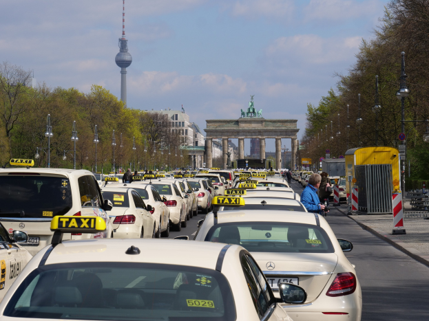 Belebte Berliner Straße mit zahlreichen parkenden Taxis, Fußgängern auf dem Gehweg, Laternen, Bäumen, Gebäuden, fernem Bogen mit Statuen, Turm und bewölktem Himmel.